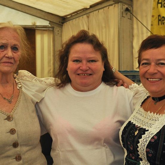 Drei Frauen stehen zusammen und lächeln für ein Foto. Die Frau in der Mitte trägt ein weißes T-Shirt, während die anderen traditionelle Kleidung tragen.