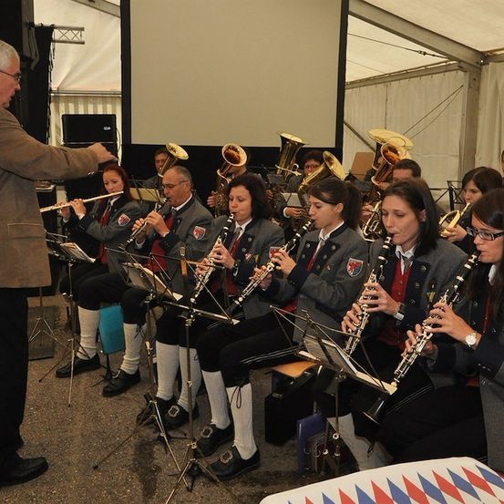 Eine Gruppe von Musikern, darunter Klarinetten und Tubas, spielt drinnen unter der Leitung eines Dirigenten. Die Musiker sitzen und stehen, mit einer großen Leinwand im Hintergrund.