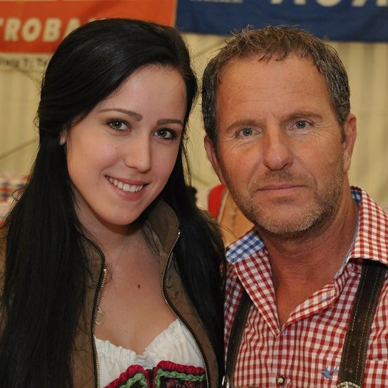 Eine lächelnde Frau mit langen schwarzen Haaren und ein Mann mit Bart und Trägerhose stehen eng beieinander, möglicherweise auf einem Festival.