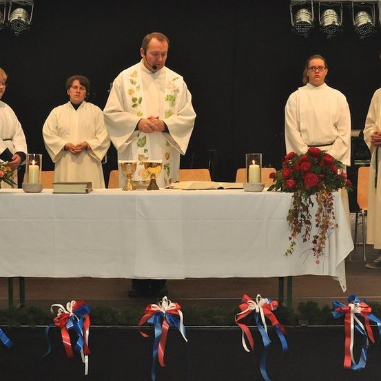 Ein Priester steht an einem Tisch mit Kerzen und Blumen, umgeben von Akolythen in weißen Gewändern auf einer Bühne.