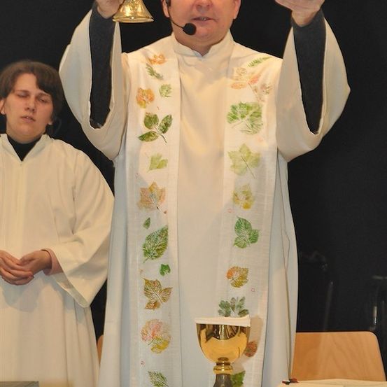 Ein Priester in einer weißen Robe mit Blattdesigns hält einen goldenen Kelch und eine klare Schüssel in einer Kirchenumgebung.