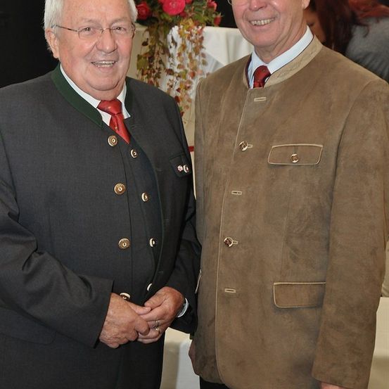 Zwei Männer in traditioneller Kleidung, lächelnd nebeneinander bei einer Veranstaltung. Dahinter eine Blumenarrangement und ein verschwommener Hintergrund.