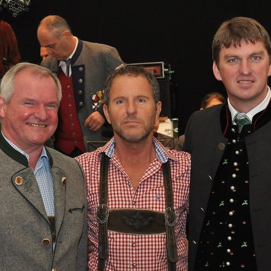 Drei Männer in traditioneller deutscher Kleidung, die lächelnd für ein Foto in einem Raum mit verschwommenem Hintergrund posieren.