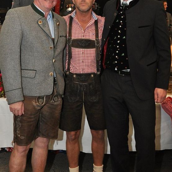 Drei Männer in traditioneller deutscher Kleidung, möglicherweise bei einem Fest, lächeln für ein Foto.