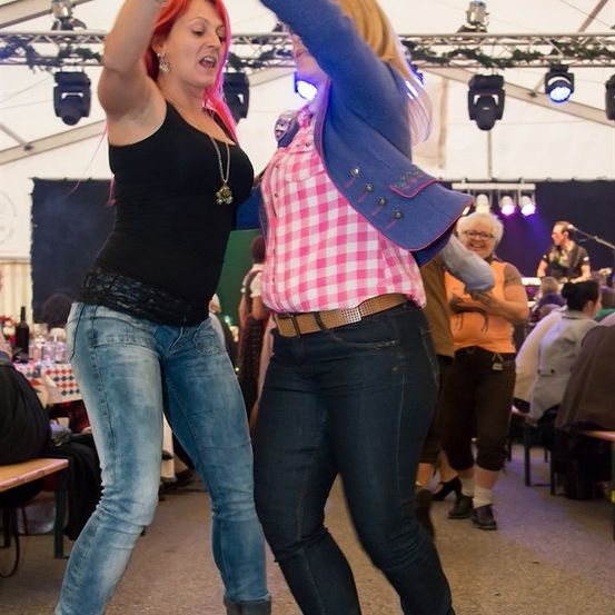 Zwei Frauen tanzen energiegeladen in einer festlichen Umgebung, mit einer Band im Hintergrund und Zuschauern an Tischen.