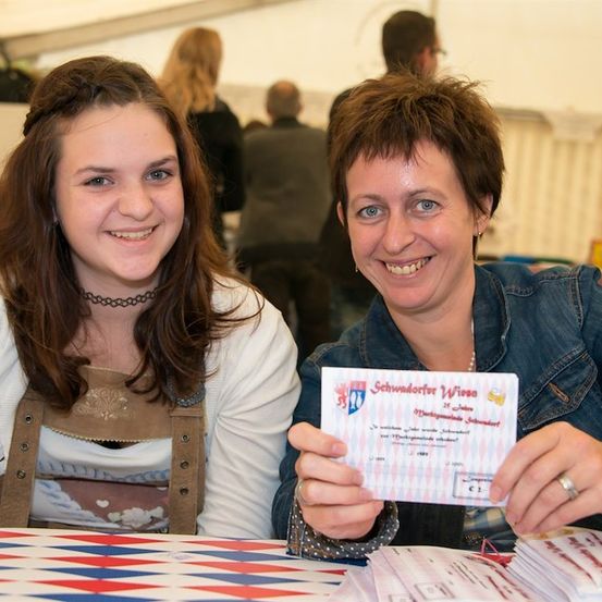 Zwei Frauen lächeln und halten ein Zertifikat in der Hand, eine trägt eine Halskette und eine Jeansjacke, die andere ein weißes Hemd mit einer braunen Weste, sitzend an einem Tisch mit einer rot, weiß und blauen Tischdecke.
