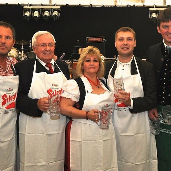 Fünf Personen in weißen Schürzen, zwei mit Bierkrügen, stehen gemeinsam auf einem Foto. Der Mann links trägt ein rot-weiß kariertes Hemd.