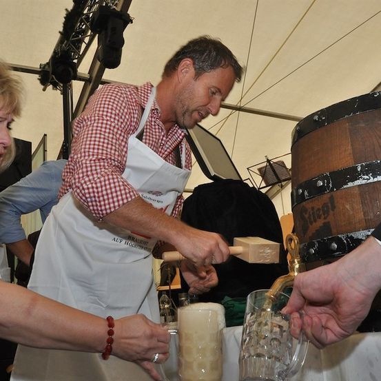 Ein Mann in einer weißen Schürze zapft Bier aus einem großen Holzfass in zwei Gläser. Eine Frau und eine weitere Person halten die Gläser. Der Schauplatz scheint ein Bierfestival zu sein.