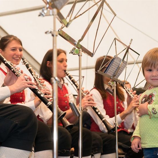 Eine Gruppe junger Musiker, in traditioneller Kleidung gekleidet, spielt Klarinetten in einem Zelt. Ein kleines Kind steht in der Nähe und beobachtet sie.