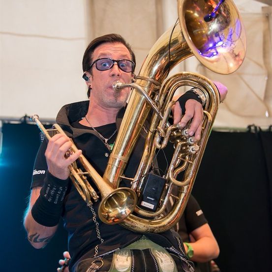 Ein Mann mit Brille und schwarzem Shirt spielt eine große Messingtuba mit einer Trompete in der anderen Hand auf der Bühne.
