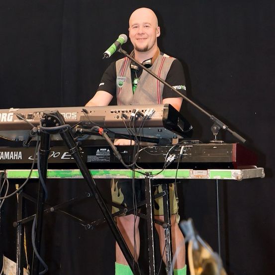 Ein kahlköpfiger Mann steht auf der Bühne, lächelt und spielt Klavier. Er trägt ein schwarzes Shirt mit grünem Logo und grüne Socken.
