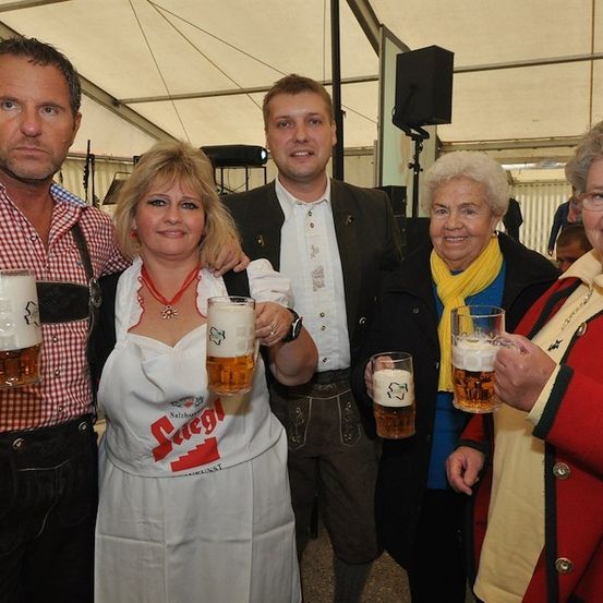 Fünf Personen in traditioneller deutscher Kleidung halten Biergläser. Die Frau in der Mitte trägt eine weiße Schürze mit dem Wort 'Suegl'. Der Mann auf der linken Seite trägt ein kariertes Hemd und eine Hose mit Aufhängung. Die Frau auf der rechten Seite trägt einen roten Mantel.