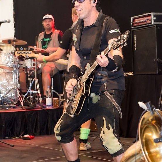 Ein Mann in einem schwarzen Shirt mit Logo spielt eine E-Gitarre auf der Bühne. Hinter ihm spielt ein anderer Mann Schlagzeug.