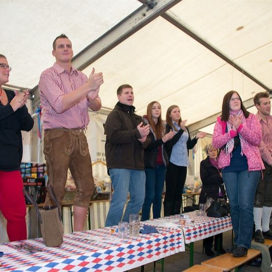 Eine Gruppe von Menschen steht und klatscht in einem Zelt, wahrscheinlich bei einem Festival. Ein Mann in einem karierten Hemd gibt einen Daumen hoch.