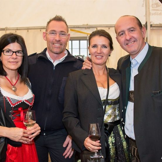 Vier Menschen lächeln und posieren für ein Foto. Die Frau links trägt ein traditionelles deutsches Kleid und hält ein Weinglas. Der Mann neben ihr trägt eine schwarze Jacke. Die Frau in der Mitte hält ein Weinglas und trägt ein grünes Kleid. Der Mann rechts trägt einen Anzug und eine grüne Weste.