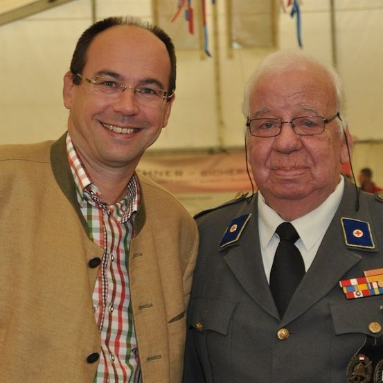 Zwei Männer nebeneinander, einer in Uniform mit Medaillen und der andere in einem lässigen Outfit, beide lächeln in die Kamera.