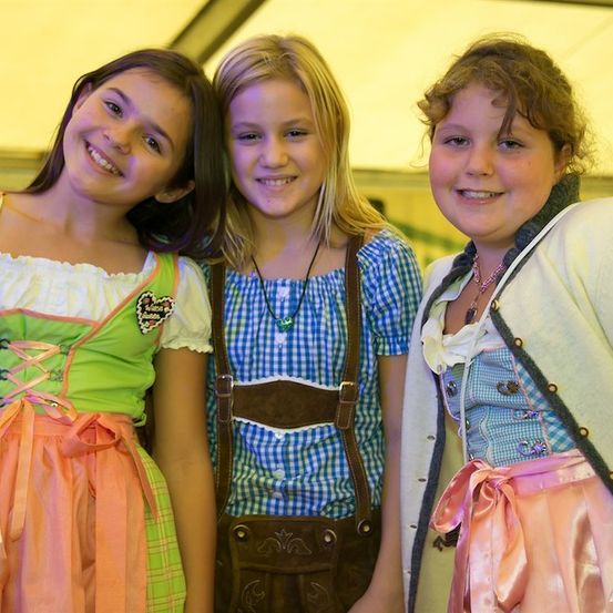 Drei junge Mädchen in traditioneller bayerischer Kleidung, lächelnd und posierend für ein Foto. Das Mädchen auf der linken Seite trägt ein grün-pinkes Kleid, die mittlere ein blau-weißes Oberteil mit braunen Trägern und das Mädchen auf der rechten Seite ein weiß-pinkes Kleid mit einer Halskette.