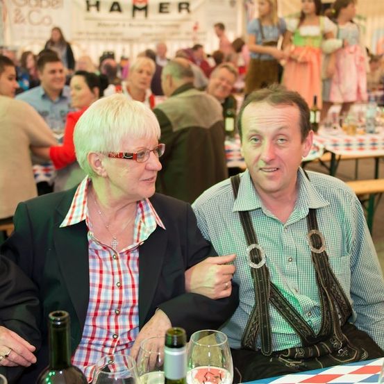 Ein älteres Paar, gekleidet in traditioneller deutscher Kleidung, sitzt an einem Tisch mit Weinflaschen und Gläsern. Sie lächeln und schauen in die Kamera. Hinter ihnen sitzen andere Leute an Tischen, und einige stehen. Im Hintergrund befinden sich Banner mit Text.