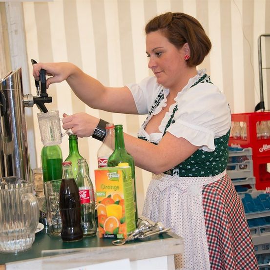 Eine Frau in einem traditionellen bayerischen Kleid schenkt ein Getränk aus einem Zapfhahn in ein Glas. Sie ist von Flaschen und einem Krug umgeben.