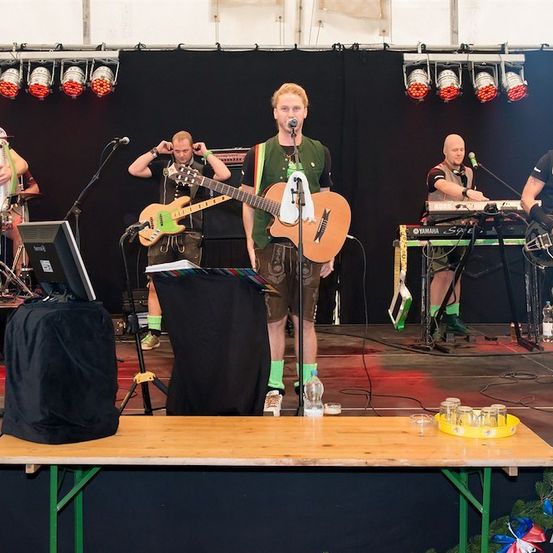 Eine Band spielt auf der Bühne mit verschiedenen Instrumenten, darunter Gitarren, Keyboards und einem Akkordeon. Der Leadsänger hält eine Gitarre und ein Mikrofon.