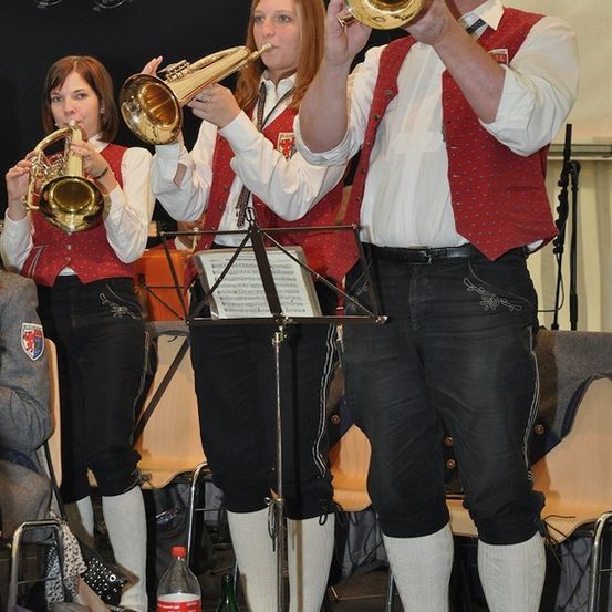 Drei Musiker in traditioneller Kleidung spielen Blechblasinstrumente auf einer Bühne, mit einem Notenständer vor ihnen und einer Flasche Coca-Cola auf dem Boden.