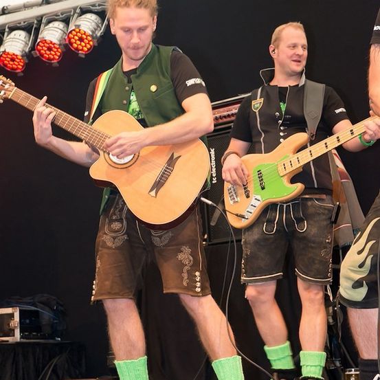 Zwei Männer auf der Bühne, einer spielt Gitarre und der andere einen grünen Bass, beide tragen grüne Socken und traditionelle deutsche Shorts.