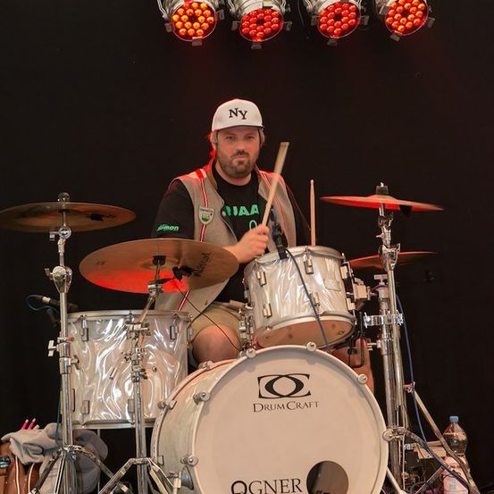 Ein Mann spielt auf der Bühne ein DrumCraft-Drum-Set. Er trägt eine weiße Kappe und eine reflektierende Weste. Mehrere Lichter sind auf der Bühne.