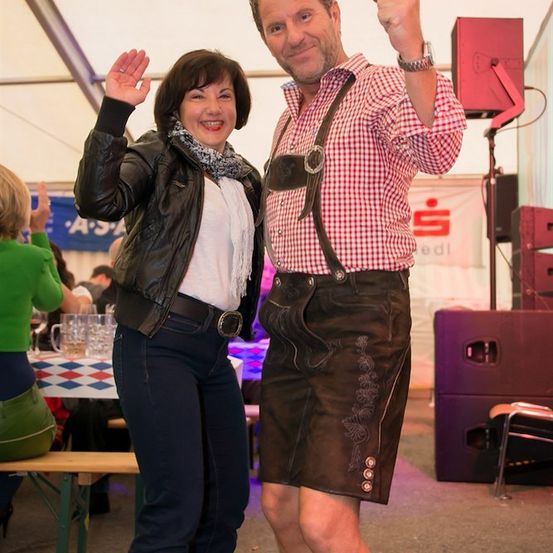 Ein Mann und eine Frau stehen bei einem Event, lächeln und winken. Der Mann trägt traditionelle bayerische Kleidung, während die Frau eine Lederjacke und Jeans trägt. Hinter ihnen steht ein Tisch mit Gläsern und einem Banner.