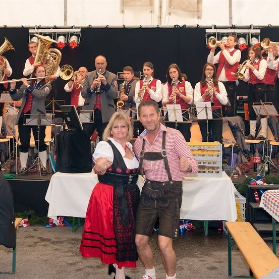 Ein Mann und eine Frau in traditioneller bayerischer Kleidung stehen vor einer Bühne, auf der ein Blasorchester spielt. Das Orchester spielt verschiedene Instrumente, und das Publikum sitzt auf Bänken.
