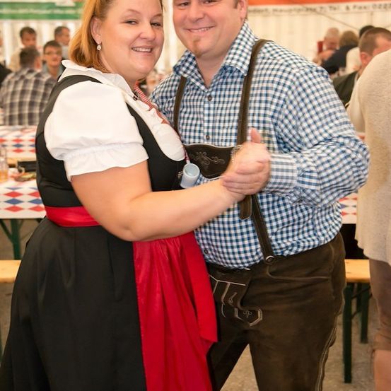 Ein Mann und eine Frau in traditioneller deutscher Kleidung tanzen bei einer Veranstaltung im Freien. Der Mann trägt ein kariertes Hemd und eine Hose mit Aufhängung, während die Frau ein schwarzes und rotes Kleid mit einem roten Gürtel trägt. Sie lächeln und scheinen sich zu amüsieren.