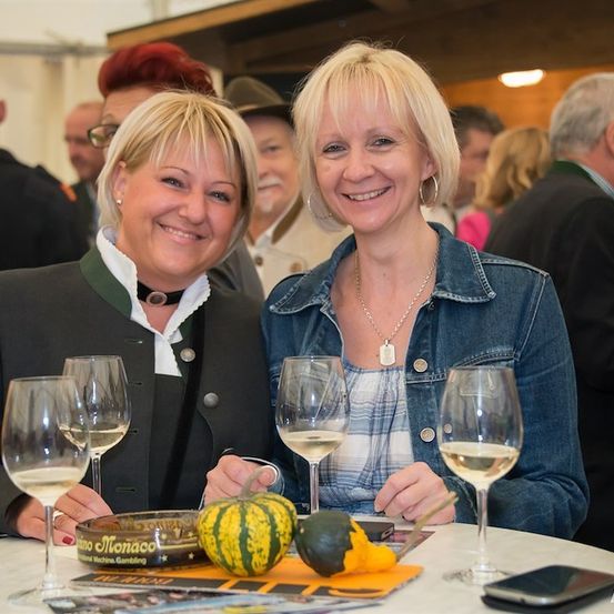 Zwei Frauen sitzen an einem Tisch mit Weingläsern und einer Dose Snacks und lächeln in die Kamera. Hinter ihnen stehen eine Gruppe von Menschen und unterhalten sich.