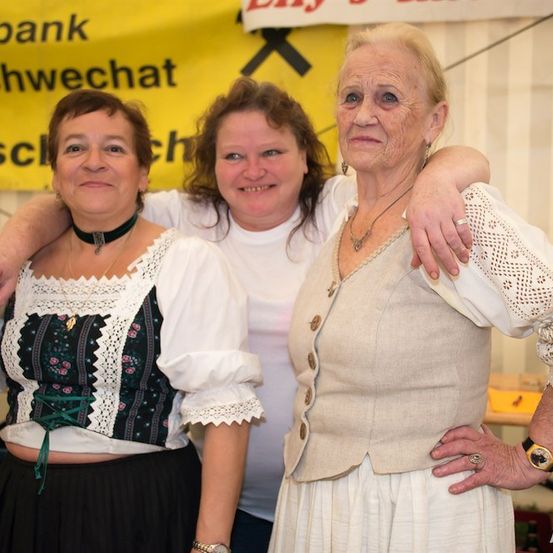 Drei Frauen stehen lächelnd zusammen, eine in traditioneller Kleidung und eine mit einer Uhr. Hinter ihnen ist ein gelber Banner zu sehen.