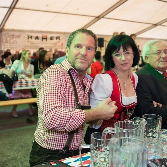 Drei Personen sitzen an einem Tisch mit Biergläsern und Krügen, möglicherweise bei einem Festival. Der Mann trägt ein kariertes Hemd und eine Hose mit Aufhängung, die Frau ein traditionelles Kleid. Der Mann rechts trägt einen Anzug und eine Krawatte.