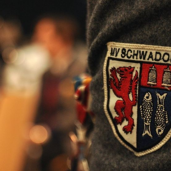 Ein Nahaufnahme eines grauen Jacken mit einem roten Löwenwappen und zwei Fischen, beschriftet 'NV SCHWADORF'. Der Hintergrund ist unscharf mit einer sitzenden Person.