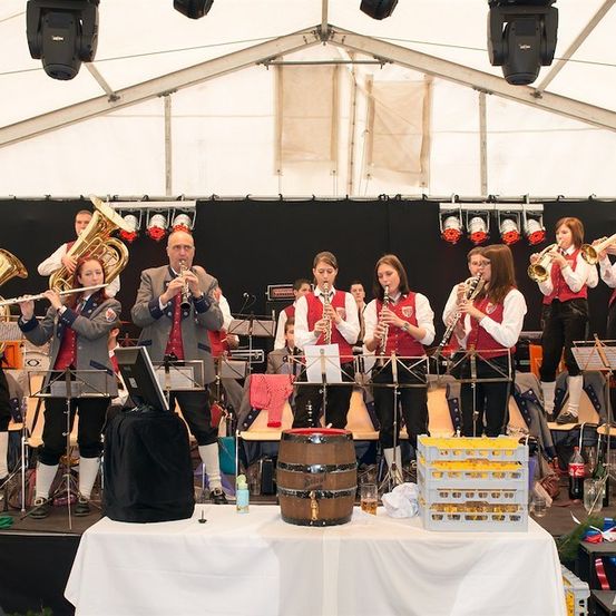Ein Blasorchester spielt auf der Bühne mit verschiedenen Instrumenten, darunter Tuben und Trompeten, unter einem Zelt mit Beleuchtungsgeräten.