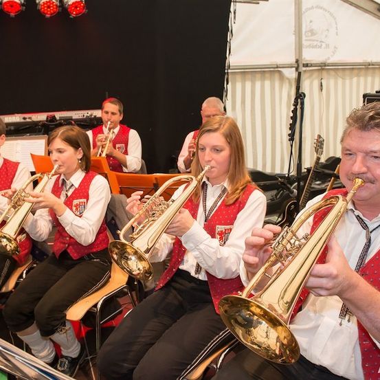 Eine Gruppe von Musikern in roten Westen und weißen Hemden spielt Trompeten in einem Zelt. Der Mann auf der rechten Seite trägt eine rote Krawatte mit Logo.