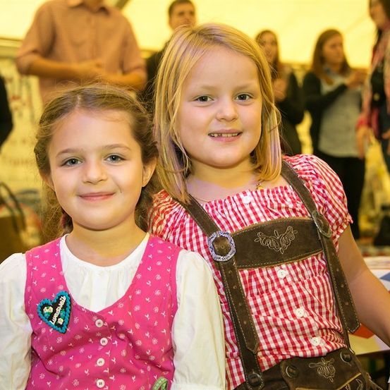 Zwei junge Mädchen in traditioneller deutscher Kleidung lächeln für ein Foto mit einem verblassten Hintergrund von Menschen bei einer Veranstaltung.