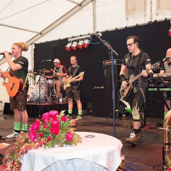 Eine Band spielt auf der Bühne mit Gitarren, einem Keyboard und einer Trompete. Der Leadsänger hält ein Mikrofon, während andere Instrumente spielen. Die Bühne ist mit Blumen dekoriert und hat einen schwarzen Hintergrund.