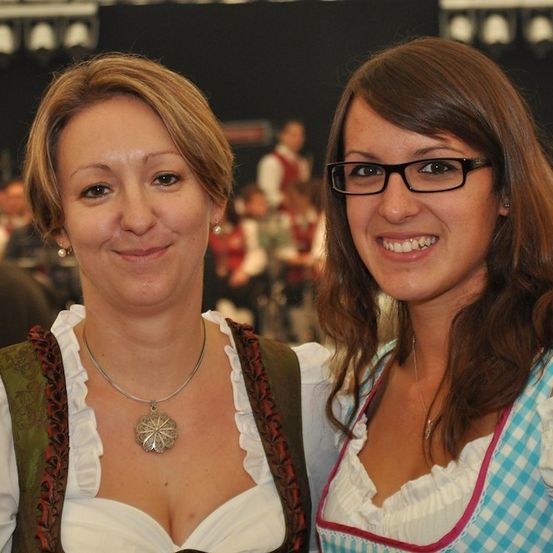Zwei Frauen in traditioneller deutscher Kleidung, eine mit einer silbernen Anhängerkette, lächeln und posieren für ein Foto bei einer Veranstaltung.