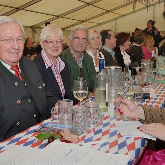 Eine Gruppe von Menschen sitzt an einem Tisch in einem Zelt, mit Weinläsern, Flaschen und einer Frau, die auf ein Stück Papier schreibt.