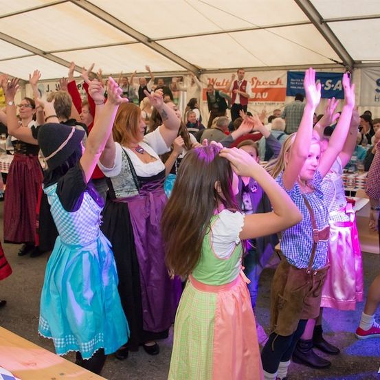 Eine Gruppe von Menschen, darunter Frauen in traditionellen Kostümen und Kinder, tanzt in einem Zelt mit einem Banner im Hintergrund.