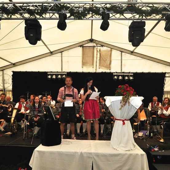 Ein Mann und eine Frau stehen auf einer Bühne vor einer Band, mit einem Strauß roter Rosen auf einem Tisch. Der Mann hält ein Stück Papier und die Frau hält ein Mikrofon. Die Band spielt hinter ihnen.