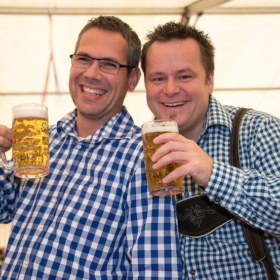 Zwei Männer in blau-weiß karierten Hemden halten Gläser mit Bier in der Hand, lächeln und posieren für ein Foto.