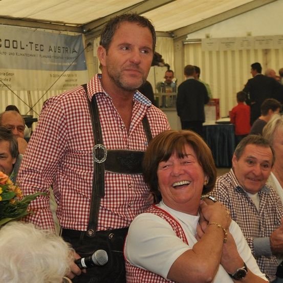Ein Mann in traditioneller bayerischer Kleidung steht neben einer lächelnden Frau und posiert für ein Foto bei einer Veranstaltung. Dahinter ist eine Menschenmenge zu sehen, und ein Banner mit Text hängt über ihnen.