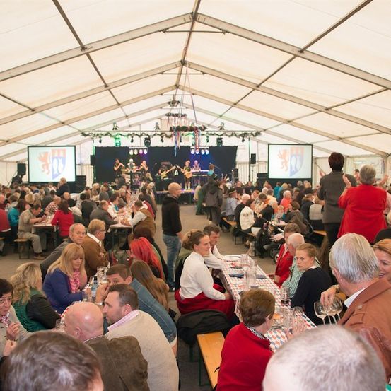 Eine große Gruppe von Menschen sitzt in einem Zelt an Tischen und genießt Essen und Getränke. Eine Band spielt auf der Bühne mit zwei großen Bildschirmen im Hintergrund.