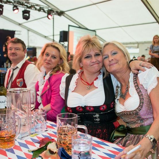 Zwei Frauen in traditioneller deutscher Kleidung lächeln und posieren für ein Foto bei einer festlichen Veranstaltung. Sie sitzen an einem Tisch mit Gläsern Bier und einer Flasche Wein. Im Hintergrund genießen andere Menschen die Veranstaltung unter einem Zelt.