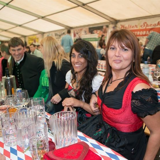 Zwei Frauen in traditioneller Kleidung sitzen an einem Tisch mit Gläsern und Flaschen und lächeln für ein Foto in einem überfüllten Zelt.