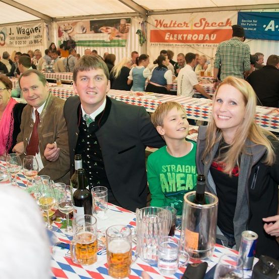 Eine Familie mit vier Personen, zwei Erwachsenen und zwei Kindern, sitzt an einem Tisch in einem Zelt, lächelt und posiert für ein Foto. Der Tisch ist mit Gläsern, Flaschen und Essen gedeckt. Hinter ihnen sitzen andere Leute an Tischen, und eine Fahne hängt an der Wand.