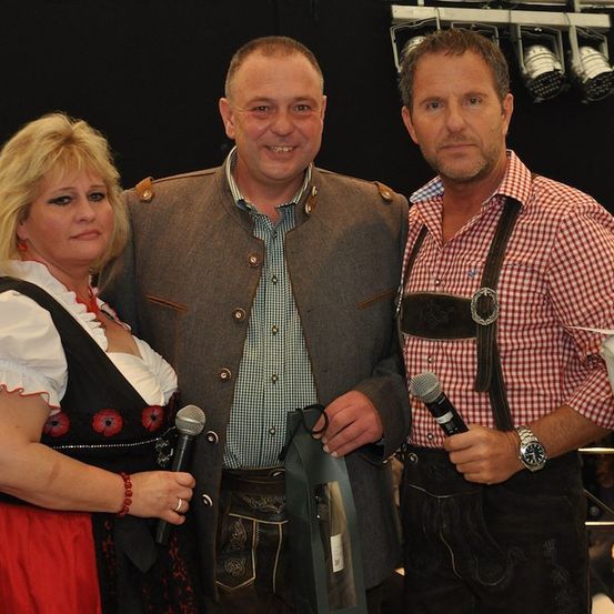 Drei Menschen in traditioneller deutscher Kleidung stehen zusammen und lächeln für die Kamera. Die Frau hält ein Mikrofon und die beiden Männer ebenfalls.