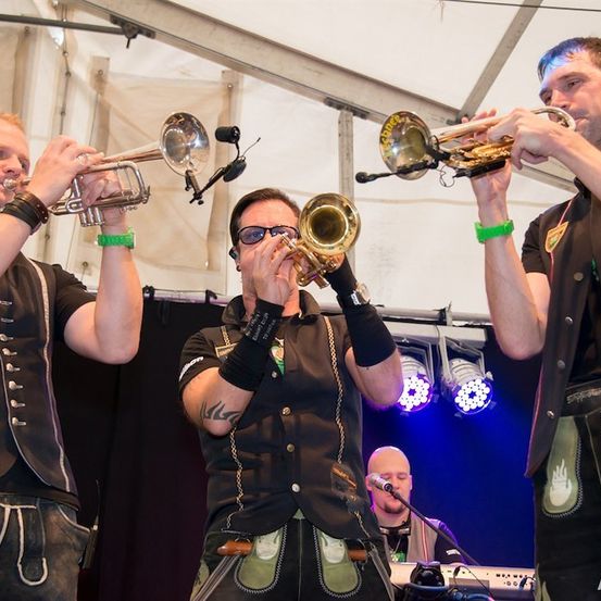 Drei Männer in traditioneller deutscher Kleidung spielen Trompeten auf der Bühne unter einem Zelt. Der Mann in der Mitte trägt eine Sonnenbrille und eine schwarze Weste. Ein Keyboarder steht hinter ihnen.